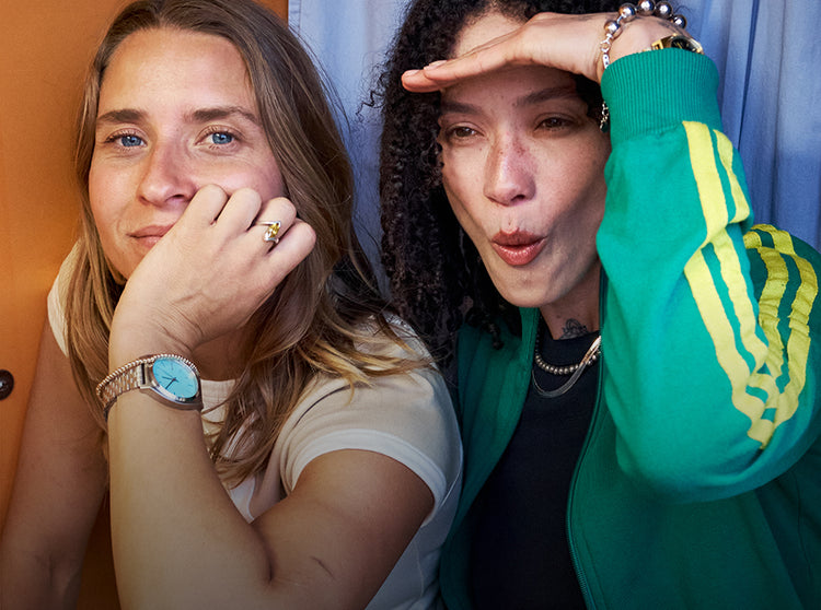 Two women, one blonde with blue eyes and a watch, the other with curly hair in a green and yellow striped jacket, looking into the distance.