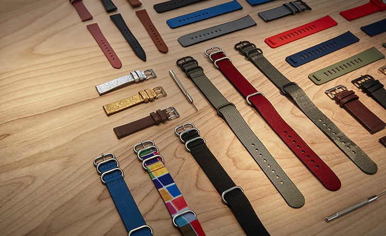 A collection of diverse watch bands in various colors, materials, and styles, including leather, silicone, and NATO straps, displayed on a wooden surface with watch tools.