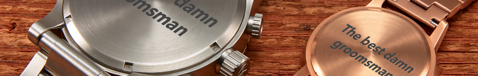 Close-up of two engraved groomsman watches, one silver with 'damn groomsman' and one rose gold with 'The best damn groomsman', on a wooden surface.