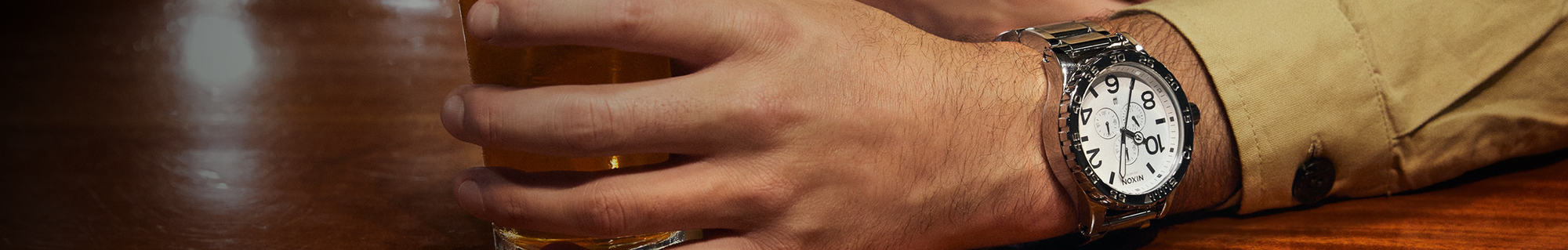 A person's arm wearing a silver Nixon chronograph watch with a white face and black numerals, holding a glass of beer on a wooden table.