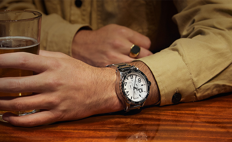 Silver Nixon chronograph watch with white dial on a man's wrist, holding a glass of beer.
