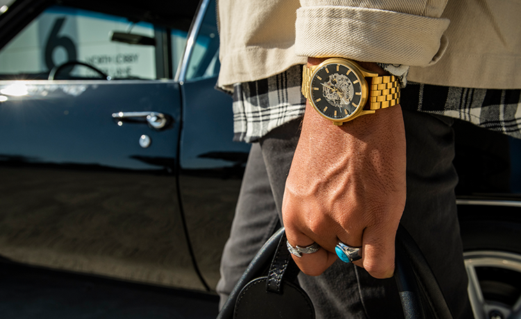 A person wearing a gold Nixon Spectra automatic watch with a visible movement, rings, and a plaid shirt, standing next to a classic black car.