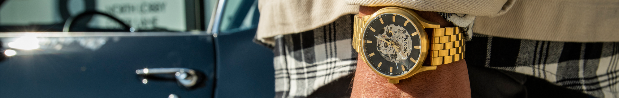 Gold Nixon Spectra automatic watch with a black skeleton dial on a person's wrist, with a blurred car in the background.