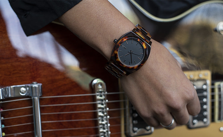 A person's wrist wearing a dark-faced watch with a tortoiseshell-patterned band, resting on a brown electric guitar.
