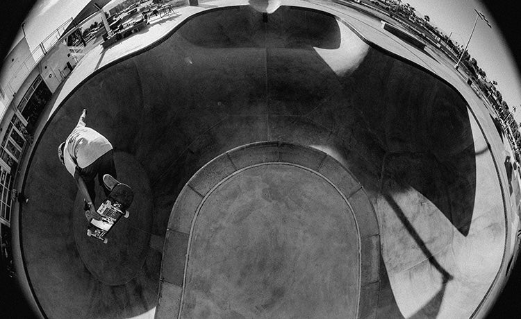 Black and white fisheye view of skateboarder Zach Miller riding in a concrete skate park bowl.