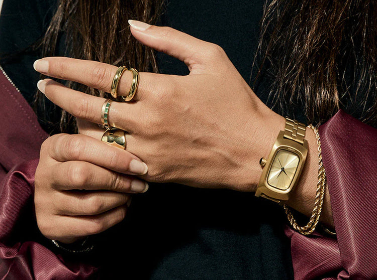 Woman with gold jewelry and a red jacket wears a gold Nixon Frankie analog watch