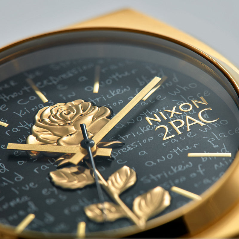 Close-up of a gold Nixon 2PAC watch with a black face, featuring a raised gold rose and leaves, and handwritten text.