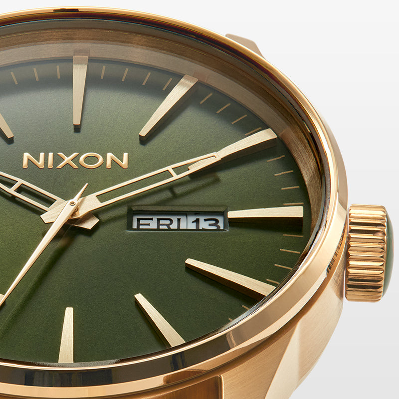Close-up of a Nixon watch with a green face, gold hands, and rose gold casing, showing "FRI 13" in the date window.