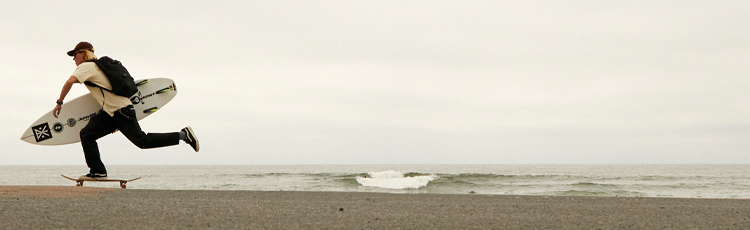 Young man with backpack and surfboard running on a skateboard along a cloudy beach.