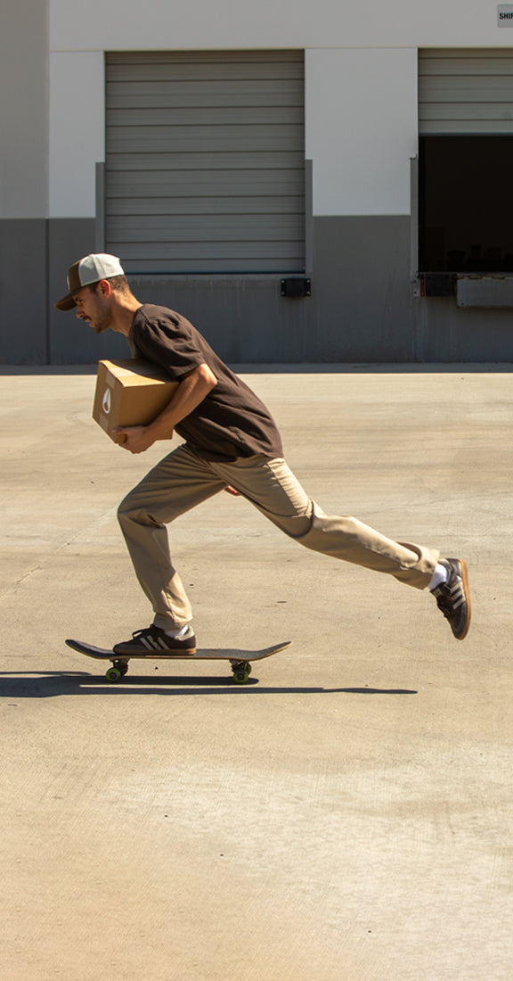 A man in a brown t-shirt and khaki pants rides a skateboard while carrying a cardboard box in an outdoor shipping lot.
