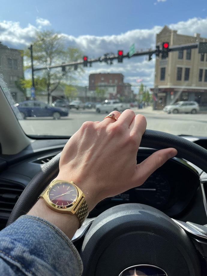 Woman driving in a city shows off her Nixon Time Teller analog watch with her hand on the steering wheel.